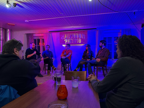 A panel discussion in a cozy, warmly lit venue with wooden walls and ceiling. The conversation is led by Paul Hudson and Mikaela Caron, hosts of the Swift over Coffee podcast. The panelists include Daniel Devesa, Klemens Strasser, and Rob Whitaker. Two audience members are seated in the foreground at a wooden table with drinks.