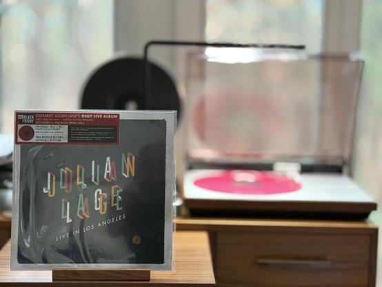 Julian Lage - Live in Los Angeles LP.
Julian’s name, on a black background with a grey edge, diagonally in multiple, 3-D colors.
The pink LP plays on a vintage, restored Bang & Olufsen Beogram 4000 linear-tracking turntable to the right.