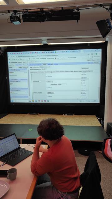 Im Vordergrund eine Person mit rotem Oberteil, am Laptop sitzend. Im Hintergrund eine Beamer-Projektion mit OpenRefine für die Arbeit in Wikidata
