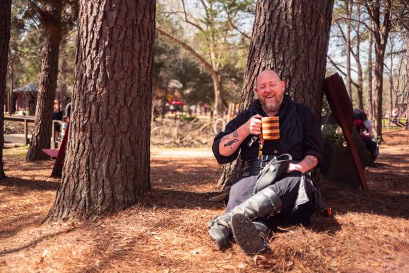 A person dressed in medieval-style attire is sitting against a large tree trunk, holding a wooden mug and smiling. They are in a wooded area, with sunlight filtering through the trees. The ground is covered in pine needles, and other people can be seen in the background, suggesting a festive outdoor event.