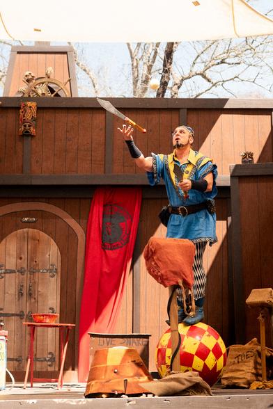 A performer dressed in a blue and yellow outfit is balancing on a large red and yellow checkered ball while juggling a knife and ball. He stands in front of a wooden backdrop with a skeleton at the top left steering a ship's wheel. A red curtain hangs behind him, and the area is decorated with various props and a drum.