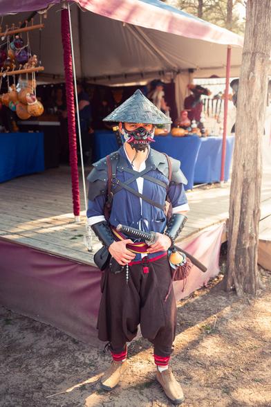 A person dressed in a samurai-inspired costume stands outdoors next to a wooden platform. They are wearing a blue tunic, dark armor pieces, a hat, and a face mask resembling traditional samurai attire. They hold a sheathed sword and have a small decorated gourd attached to their belt. In the background, there is a tent with blue-covered tables displaying colorful items.