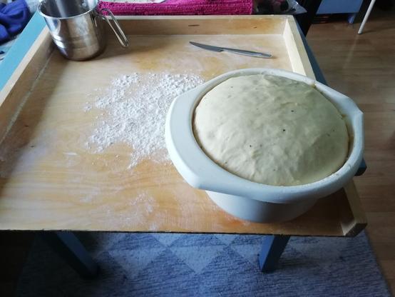 Pöydällä on puinen leivintaso. Sen päällä on valkoinen muovinen kulho, jossa on reunojen tasalle kohonnut pullataikina.