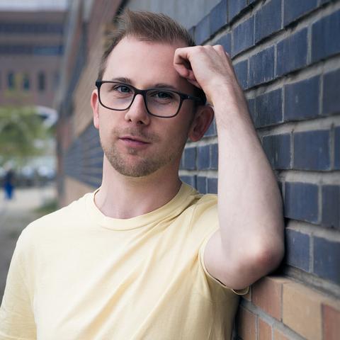 Junger Mann in gelben T-Shirt mit Brille lehnt sich an Klinkermauer.