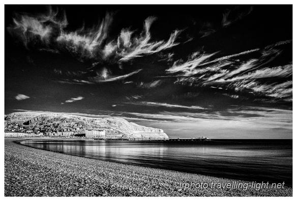 Black and white photo of calm sea lapping on a curved shingle beach. In the background is a headland with a long amusement pier in front of it. There are feathery clouds against a black sky.