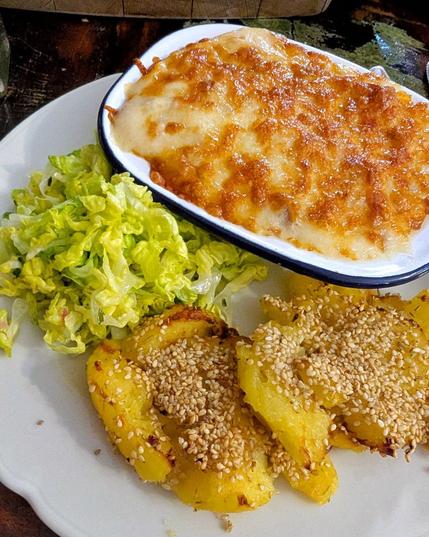White plate, smashed baked potatoes with sesame on top, green, shredded romana salad and a small pie dish filled with something that has a lot of brown cheese on top.