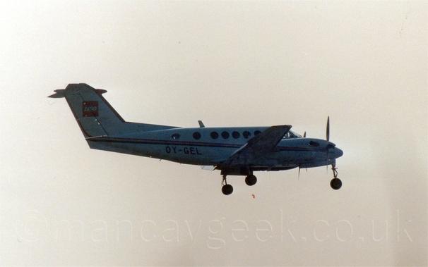 Slightly grainy and backlit side view of a white, twin propellor-engined BizProp flying from left to right at a low altitude, with undercarriage lowered and flaps extended behind the wings, suggesting it is about to land.
There is a red and black stripe running along the body, as well as on the engine cowling, and the registration "OY-GEL" on the lower rear fuselage in black.
There is a red square with a white "Lego" logo on the tail.
Bright but hazy sky, with a slight orange tinge, fills the rest of the frame.