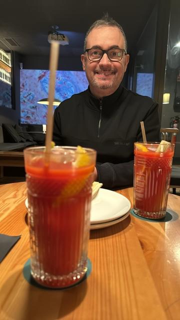 Un hombre con gafas y sudadera negra sonríe mientras está sentado en un bar con ambiente moderno y luces tenues. En primer plano, dos cócteles de color rojo intenso con hielo y rodajas de cítricos están sobre la mesa de madera, acompañados de pajitas de bambú. Al fondo, se proyecta una imagen abstracta en la pared.