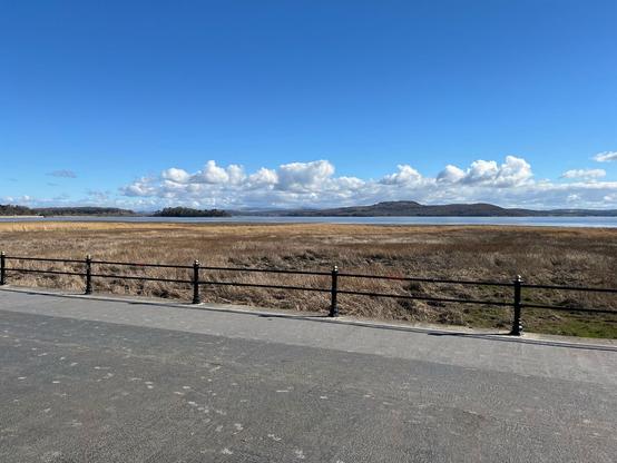 A scenic view of a grassy landscape leading to a calm body of water, under a clear blue sky with scattered clouds. A black railing lines the foreground, while distant hills are visible across the water.