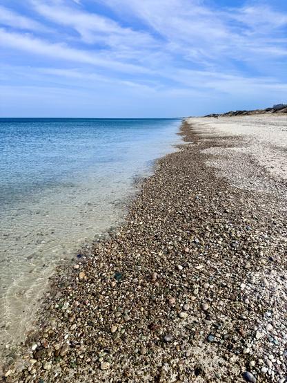 Photo looking along the shoreline of an ocean beach. Wet pebbles along beach and no surf or waves with this flat calm water. The shoreline goes to the horizon. Some green foliage seen on land at the horizon. Mostly white streaky clouds with blue sky between them.