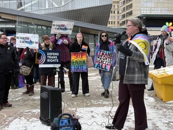 A group of people with signs and a speaker wearing a pride flag.