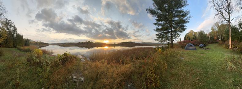 A wide panorama at sunset in Northern Ontario in early October. The sky is mostly cloudy, with a patch of blue sky behind a very tall pine tree.  In the foreground is long grass, and beyond that is a smooth, glassy river.