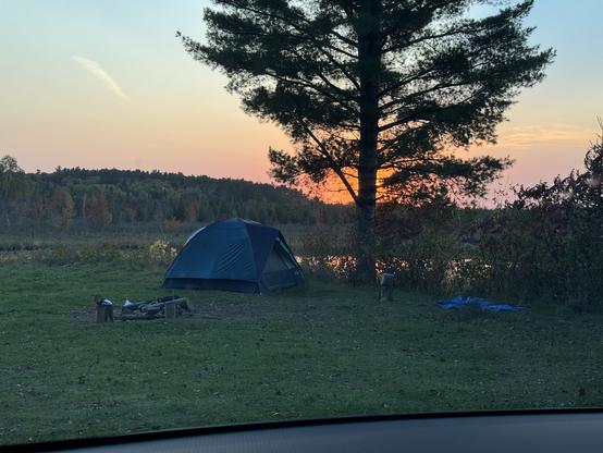 View of my campsite in the wilds of Northern Ontario at sunset. The sky above where the sun has set is almost pumpkin-orange behind the tall pine beside my tent.    The top of the photo captures the remaining blue sky before it gets dark and the coyotes start howling.