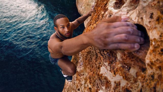 World-class climber Kai Lightner as he scales the craggy seaside cliffs of Mallorca, Spain. The background features a shimmering blue sea.