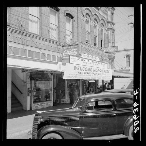 The image is a black and white photograph capturing an urban street scene from the mid-20th century. In the foreground, there's a vintage car parked on the right side of the frame with prominent features like chrome details and rounded bodywork indicative of automobiles manufactured in the 1930s or '40s.

The backdrop is occupied by various commercial establishments including a theater marquee reading "WELCOME HOP-PICKERS" along with other signs for services such as real estate, drug store, confectionery fountain, health service, and soda. The architecture of the buildings suggests an era when brick structures were prevalent in urban planning.

The signage also indicates local businesses like Cravens Drug Store and Coca-Cola advertising a "Health Service," hinting at a community centered around small business enterprises catering to daily needs. People can be seen walking on the sidewalk, suggesting pedestrian activity typical for such locales during that period.

Overall, this image portrays everyday life in what appears to be an American town with emphasis on local commerce and public gatherings highlighted by the mention of hop pickers, which is a reference to economic activities like hops agriculture common around certain regions. The photo conveys a sense of place characterized by its mix of architectural styles, business signage, and community interaction.