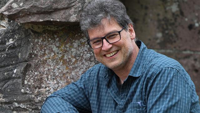 Porträt von Wilfried Röcker: Ein lächelnder Mann mittleren Alters mit grau melierten Haaren und Brille. Er sitzt vor einem Hintergrund aus grobem Mauerwerk.