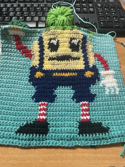 This is an image of a tapestry crochet piece depicting an onion shaped mascot wearing red and white striped stockings, black boots, Jean overalls, with red sleeves abs white hands.