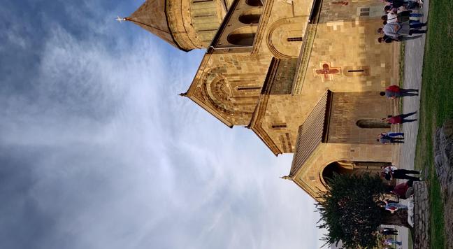 View of the cathedral from outside, with ocre sandstone and roofs at different angles, against the blue sky with a dramatic cloud.