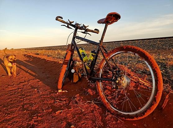 A bicycle resting on a pedal. The tyres coated in sticky red mud on a red dirt track. To the left Winnie-dog looks on.