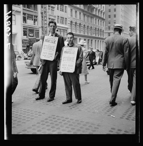 The image captures a moment from what appears to be the 1950s, showing two men holding picket signs on a busy city street. They are in front of multiple story buildings and pedestrians can be seen walking past them. The sign held by one man reads "EMPLOYEES OF PAVELLE ON STRIKE," indicating they might be part of labor actions during that period.

The image conveys the atmosphere of urban life with its black-and-white aesthetic, which gives it a historical context suggesting it is documenting social or economic conditions from past decades. There's also street signage like "PRE CANDY" and storefronts visible in the background, adding to the setting as being downtown New York City.

The photo carries an air of candidness capturing real-life events without staging or embellishment typical for contemporary photos. The presence of labor union picketing suggests a snapshot from American history involving economic struggles and worker movements during that era.