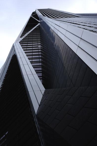 A view up a large grey building, it has long running diagonal and vertical lines with horizontal window layers inbetween. It’s reflective and dark. The sky is a grey-blue mush, very distinct from the sharp and defined building lines