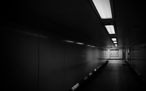A monochrome picture of a dark, empty tunnel in an underground train station. The overhead lighting is barely illuminating the darkness, the perspective is offset but drawing the eye to the brightly lit passageway that intersects in the distance.
