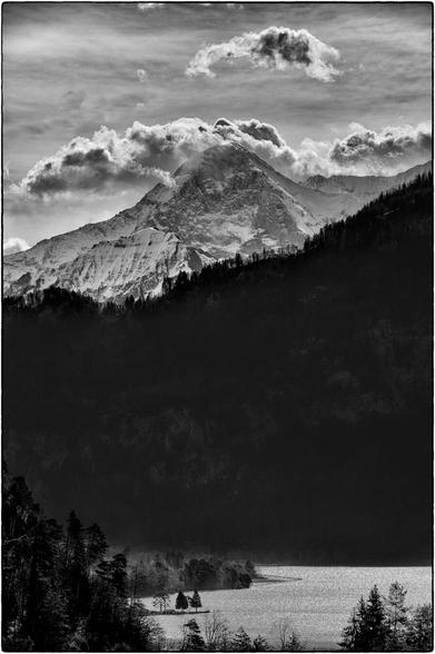 Schwarzweissfotografie im Hochformat gedrittelt: im oberen Drittel Berg und Wolken, in der Mitte Wald und im unteren Drittel See und Bäume