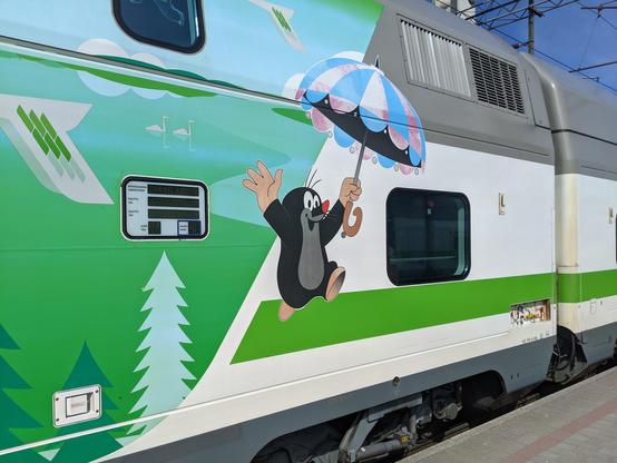 Gros plan sur une voiture de train intercités finlandais à deux niveaux. À gauche, une décoration évoquant des sapins sur fond vert, au centre une taupe souriante qui lève les bras et tient un parapluie.
