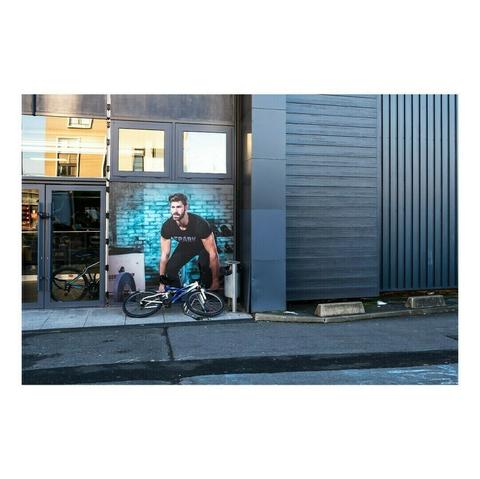Une photographie prise en extérieur montre une façade de bâtiment moderne, composée de panneaux métalliques gris et de grandes fenêtres réfléchissantes. Sur l’un des murs vitrés, un immense poster publicitaire représente un homme musclé vêtu d’un t-shirt noir, en position inclinée comme s’il était en plein effort physique dans une salle de sport. L’illusion est renforcée par la présence d’un vélo réel, posé juste devant l’affiche, donnant l’impression que l’homme en arrière-plan est en train de le tenir. Ce jeu visuel crée un effet humoristique et trompe-l’œil, où le réel et l’image imprimée semblent fusionner.