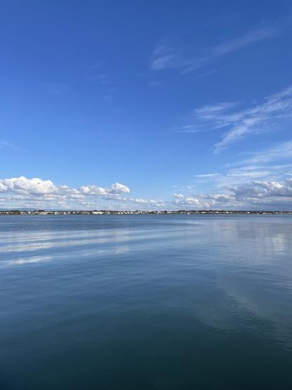 Top half of the photo is filled with blue sky with some small clouds on the horizon. The bottom half is filled with the super calm river that reflects some small houses seen in the distance. Everything is blue and very calm