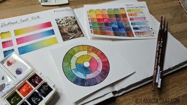 A messy desk with an open sketchbook and some bright swatches, a color circle showing the mixes is in the center, another slightly bigger mixing chart, a tiny 6 color palette all colors looking dark because the pigments are transparent and the orange being the only bright one looking pleasantly out of place. On the page is a tiny landscape showing an autumn forest that feels just as out of place as the orange due to the muted tones of the painting amidst the colorful charts.