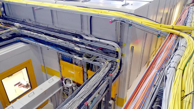 Max, das bunte Strickhorn, hat sich auf diesem Bild irgendwo versteckt. Wir schauen von oben auf Strahlenschutzeinhausungen einer Synchrotron-Messstation, zahlreiche Kabelkanäle sind zu sehen mit vielen Kabeln unterschiedlicher Farben (insbesondere die gelben Netzwerkkabel stechen ins Auge) und unterschiedlicher Dicken. Unten links ist ein graues Strahlrohr zu sehen, durch welches der Röntgenstrahl zum Experiment geleitet wird; daneben steht eine kleine Leiter, von wo aus sich bestimmt gut ein weiteres Kabel verlegen lässt.
Ivryyrvpug vfg Znk wn qbeg mh frura … ?