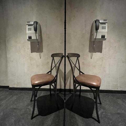 A classic, lightweight, old wooden chair next to a boxy pay phone mounted to the wall. The whole scene is mirrored in a mirror wall