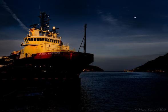 A night time photo of a harbour with a large boat on the left of the frame. The hull of the boat is dark, but the deck area is lit up. There is a row of small, dark port holes or windows on the bridge. There are two hills rising in the distance on either side of the entrance to the harbour. A dark low cloud or fog lies across that part of the scene behind the hills, with clearer skies above, a vapour trail disappearing behind the bridge of the boat from the left and a small 3/4 moon in the sky above, on the right side.