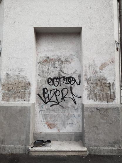 A photograph of what once was a doorway, now bricked up, and in front of it sat on a step are two pans semicolon a saucepan and a frying pan.