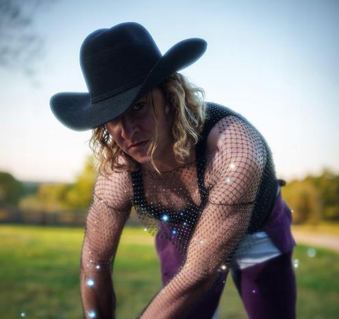 A muscular AMAB person crouches on the ground and looks directly into the camera. They are wearing a black cowboy hat, black tanktop, and a glittery fishnet shirt. In the background is a sunny field and trees.