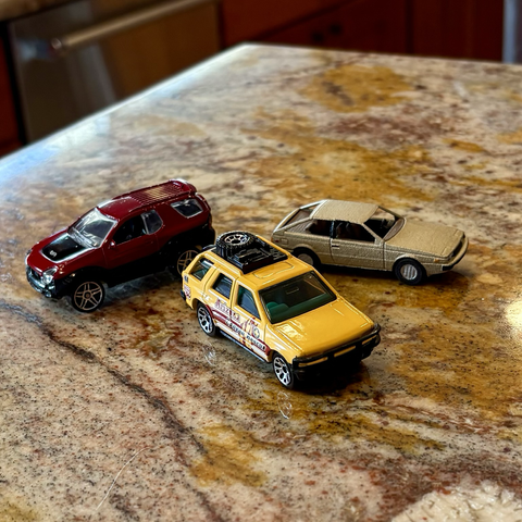 Three 1:64-scale toy Isuzu cars sit on a brown granite countertop. From left to right: a red Hot Wheels VehiCROSS SUV; a yellow Matchbox Rodeo SUV; a gold 3D-printed Impulse coupe of my own design.