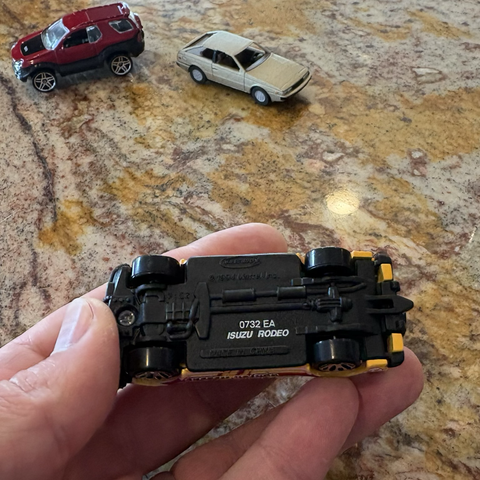A view of the underside of the yellow Matchbox Isuzu Rodeo, showing the make and model printed in white ink on the black plastic base, rather than being molded into the plastic in typical Matchbox fashion.