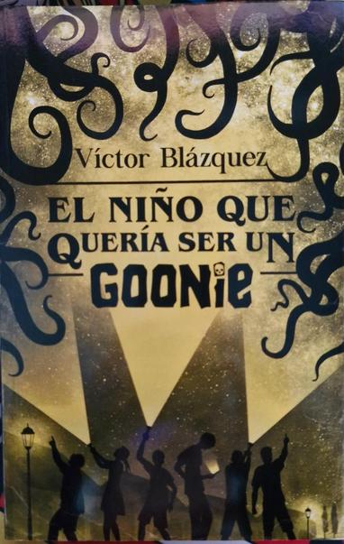 Fotografía de la portada del libro: "El niño que quería ser un goonie" de Víctor Blázquez. En ella se ven en la parte de abajo siluetas de personas, algunas con linternas iluminando el cielo y una a la cuál se le ilumina el rostro por la luz que emite la pantalla de su teléfono móvil. En la parte de arriba se ven como siluetas de ramas. Todo en colores negro y amarillo.