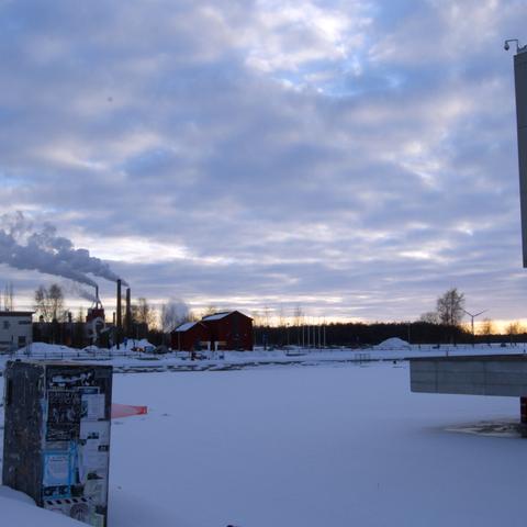 Oulu waterfront during winter, by the water near dock. Electrical box covered by posters is on the left side of the scene. The theatre building can barely be seen across the water on the right edge of the picture. In the distance in the area behind the harbour, old storehouses can be seen. In the far distance, across the bay, an industrial plant is emitting smoke.