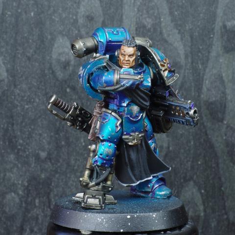 Front view of a space marine miniature. They're standing up, looking at a screen on their right arm and holding their bolter on the other. They're painted in mettalic blue. Their skin tone is olive.