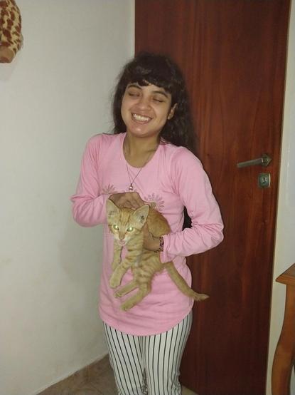 Una mujer sonriente con cabello oscuro y rizado está sosteniendo un gato anaranjado. Ella lleva una camiseta rosa y pantalones a rayas blancas y negras. El gato tiene los ojos brillantes debido al reflejo de la luz. Ambos están frente a una puerta de madera.