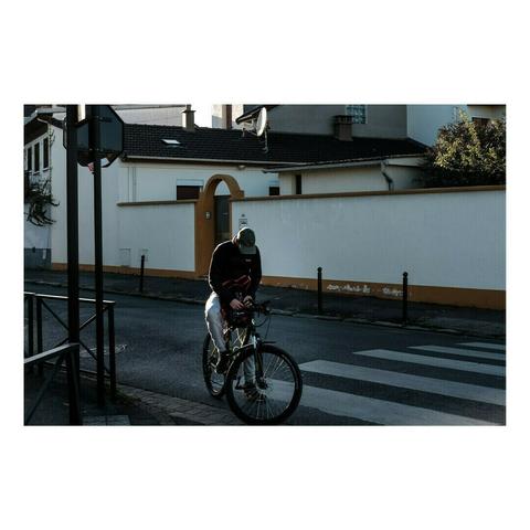 Un homme en casquette et sweat à capuche est assis sur son vélo au milieu d’un passage piéton, concentré sur son téléphone. La lumière du matin ou de la fin de journée accentue les contrastes, projetant son ombre sur l’asphalte. En arrière-plan, une maison avec un mur blanc et une porte arquée.