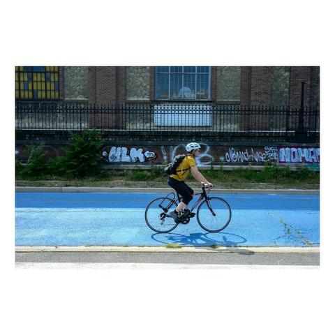 Un homme en tenue sportive et casque roule sur un vélo de course le long d’une piste cyclable peinte en bleu vif. Derrière lui, un mur recouvert de graffitis et de tags borde une ancienne usine en briques avec des fenêtres grillagées.