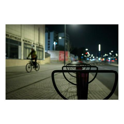 Un cycliste vêtu d’un manteau sombre traverse une avenue éclairée par des lampadaires. L’avant-plan montre des arceaux de stationnement vides, donnant une perspective répétitive et floutée. Les lumières de la ville scintillent au loin.