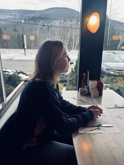 Photograph of a young woman in profile view, sitting in a booth at a diner with her hands folded on the tabletop in front of her. A winter landscape with a sparsely filled parking lot, snowy ground, leafless trees, and low rolling mountains is visible through the large plate glass windows behind her.