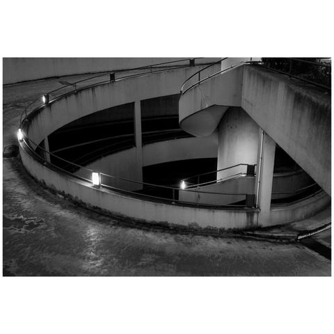 A black and white photo of a car entrance to an underground carpark during evening. The architecture is perhaps from the 70s or 80s with a lot of visible concrete. A part of the spiral road can be seen in the entire fram and on the top right corner is a part of the pedestrian access. The area is lit by lights build into the railing.