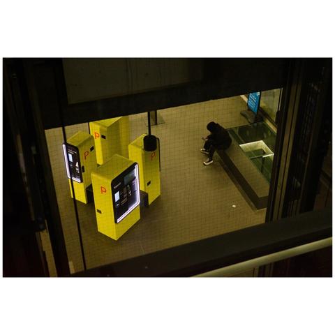 A colour photo of a subground floor photographed from above. The view is heavily framed by the architecture of the building. Four parking ticket vending machines can be seen which are painted with a vibrant yellow. A person is sitting on a bench next to the machines looking at their smartphone.