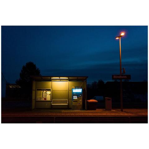 A colour photo during late sunset of a waiting shelter at a train platform. The shelter is on the left and middle of the frame and on the right is a street lamp. The differnt colour temperatures of the sky, the street lamp and the lamp in the shelter work very well together.