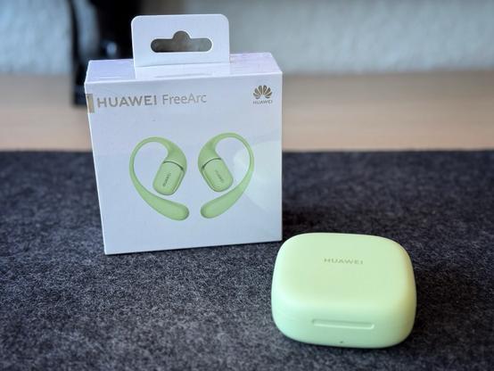 Ladecase der grünen Farbvariante der Huawei FreeArc liegt neben der Verpackung des Kopfhörers auf einer anthrazitfarbenen Schreibtischunterlage aus Filz.